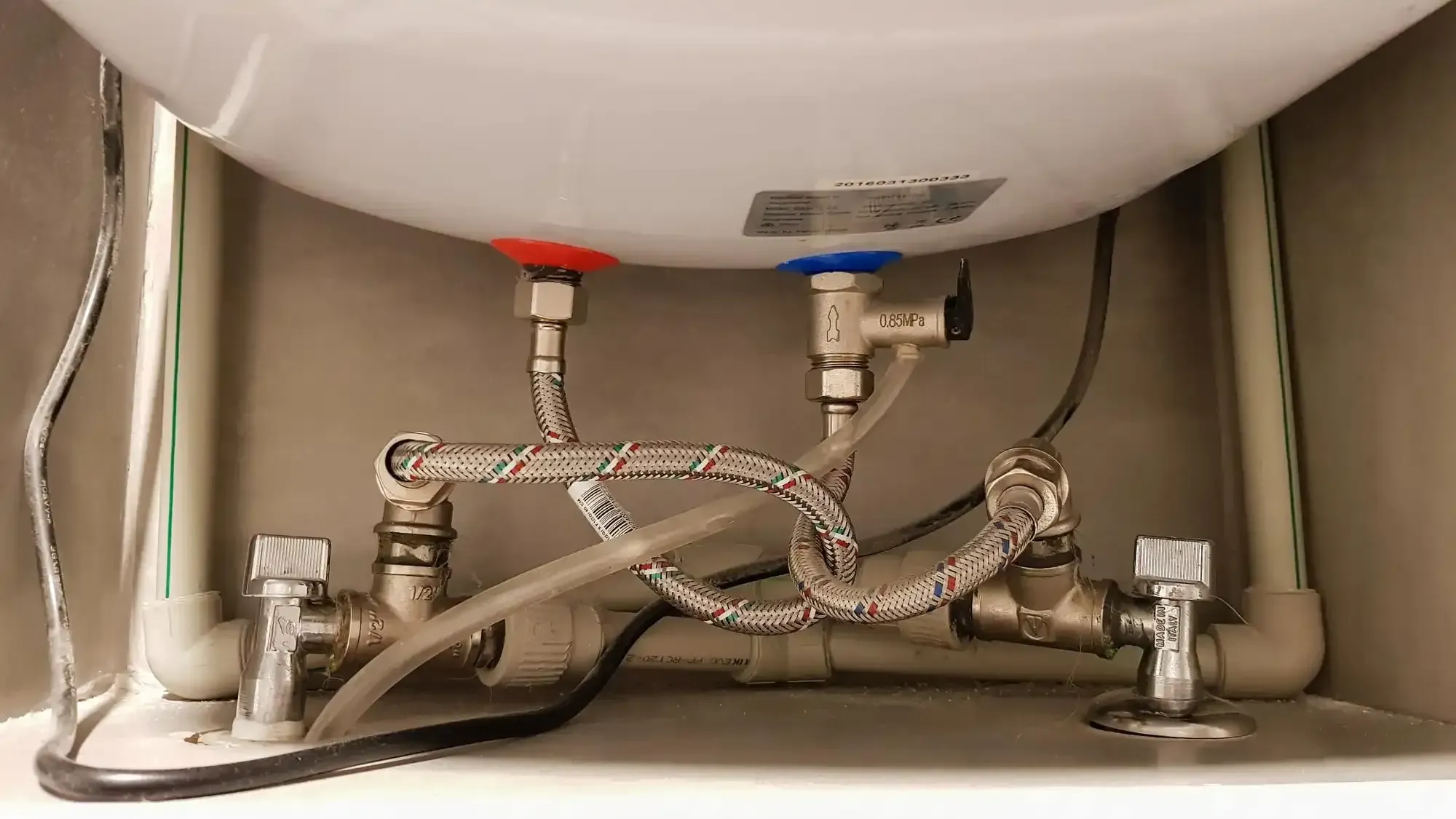 Braided supply lines and copper piping connected to a water heater inside a wall access panel.