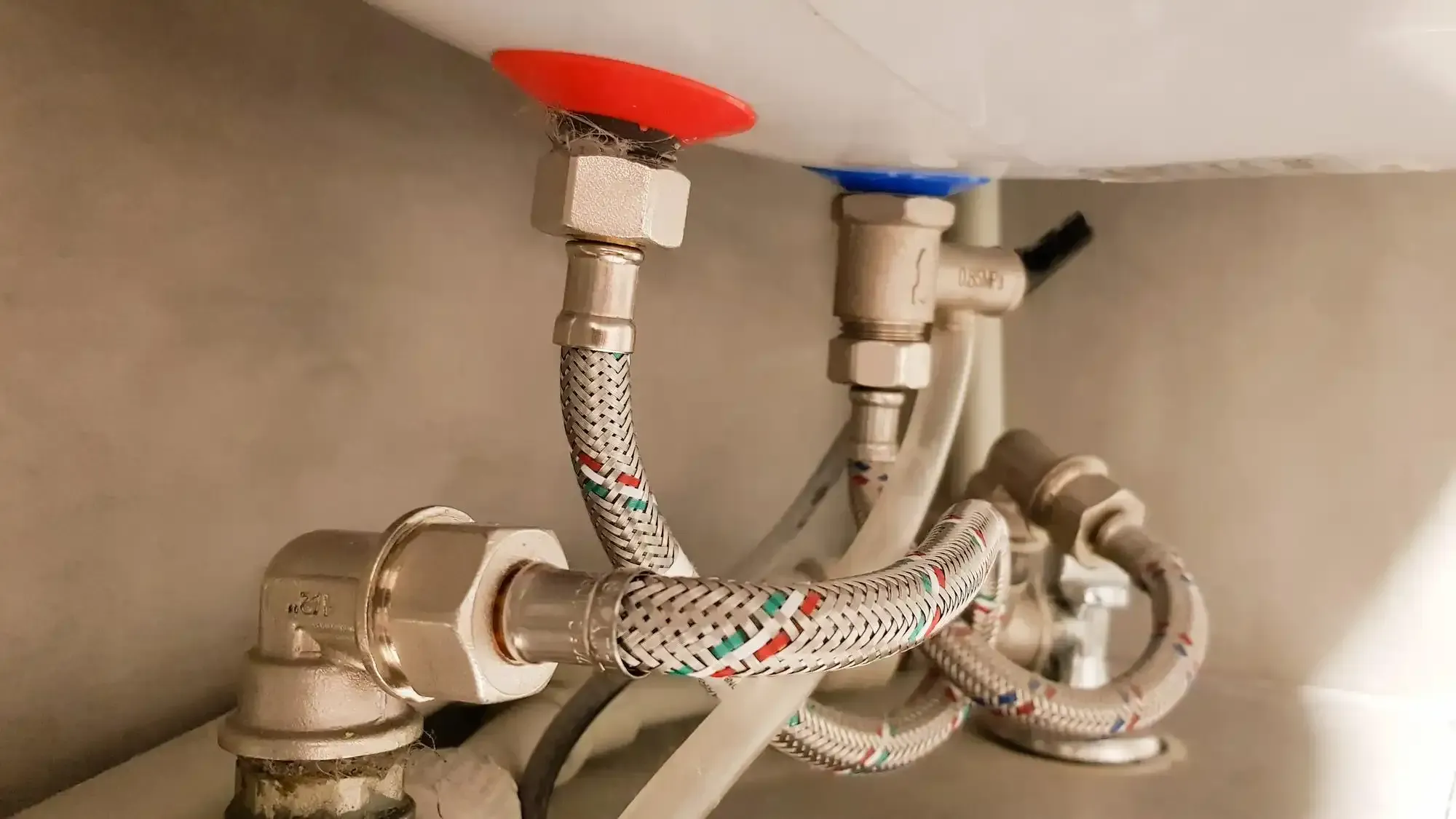 Plumbing connections beneath a white tank: red and blue caps, flexible braided hoses, and metal fittings.