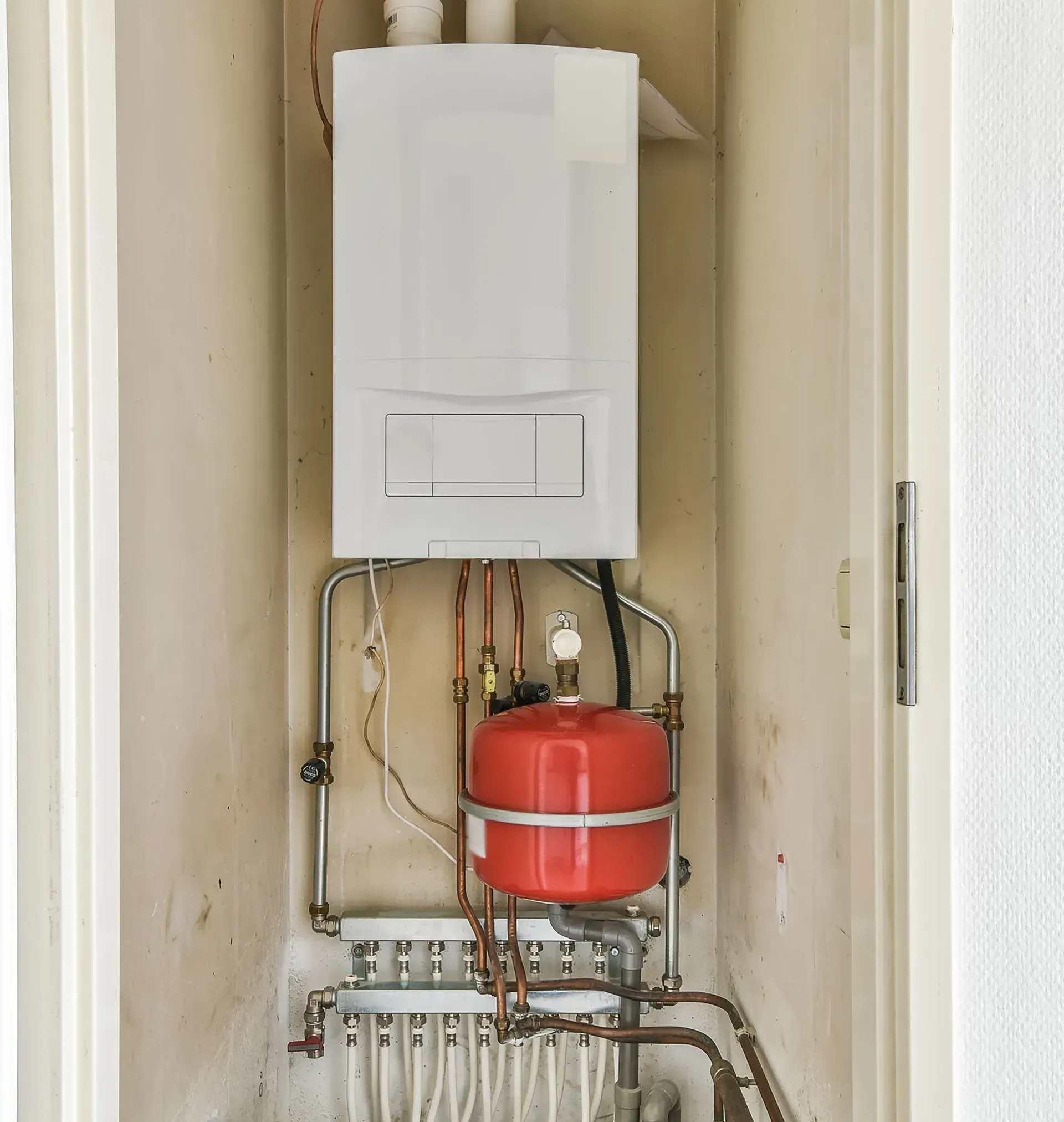 White wall-mounted boiler with red expansion tank and copper pipes in a utility closet.